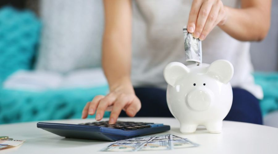 Person inserting cash into a piggy bank while using a calculator, illustrating financial savings and budgeting tips for reducing energy bills.
