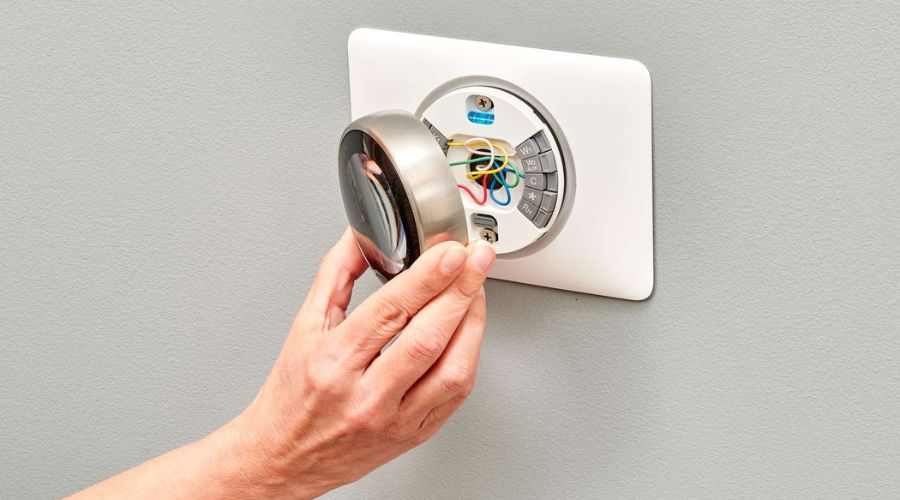 Hand adjusting a thermostat on a wall, showing wires and digital interface, relevant to HVAC maintenance and thermostat replacement discussions.