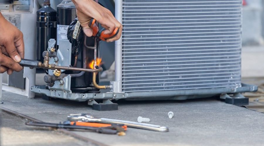Technician repairing an air conditioning unit, using tools to address a refrigerant leak, with HVAC components visible in the background.