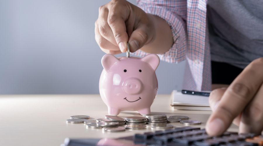 Person inserting a coin into a pink piggy bank, surrounded by coins and a calculator, symbolizing saving money while managing heating and cooling costs.